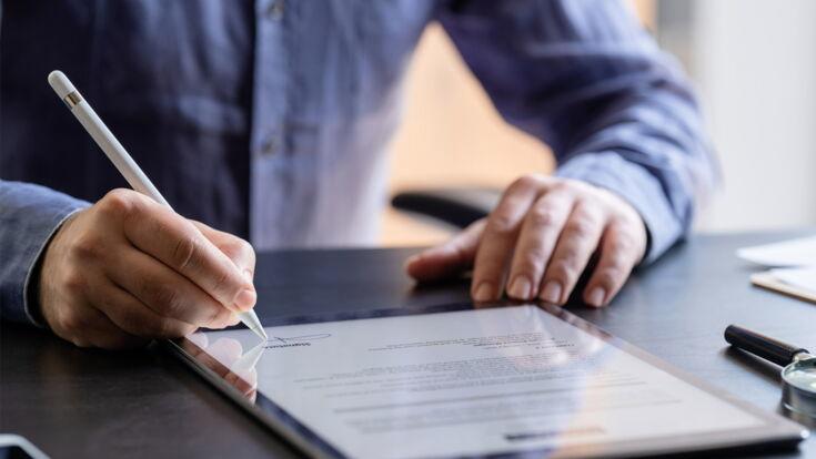 Eine Person unterzeichnet einen digitalen Vertrag auf einem Tablet mithilfe eines Stylus, während das Tablet auf einem Schreibtisch liegt. Ein Smartphone und andere Büromaterialien sind ebenfalls auf dem Tisch zu sehen.