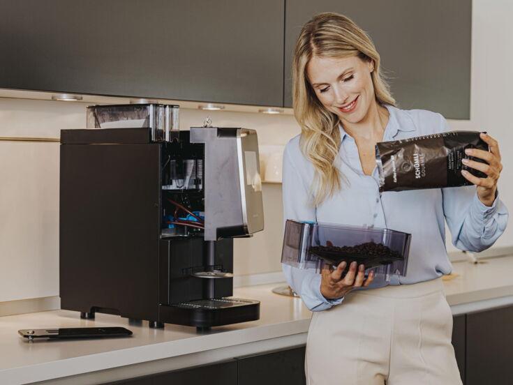 Frau steht lächelnd vor einer Küchenzeile, auf dem sich ein offener Kaffeevollautomat steht, und füllt den Produktbehälter mit Kaffeebohnen auf, den sie in der Hand hat