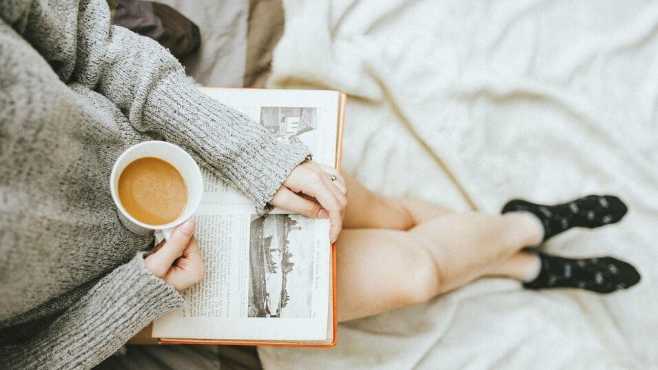 Frau liegt im Bett, liest ein Buch und trinkt schwarzen Kaffee