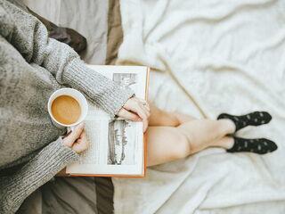 Frau liegt im Bett, liest ein Buch und trinkt schwarzen Kaffee