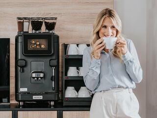 Frau lächelt, während sie eine Tasse frisch gebrühten Kaffee vor der Barista Ultima Kaffeemaschine in einem Büroumfeld hält.