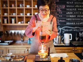 Eine asiatische Frau rührt mit einem Holzlöffel den Kaffee in einem Syphon um