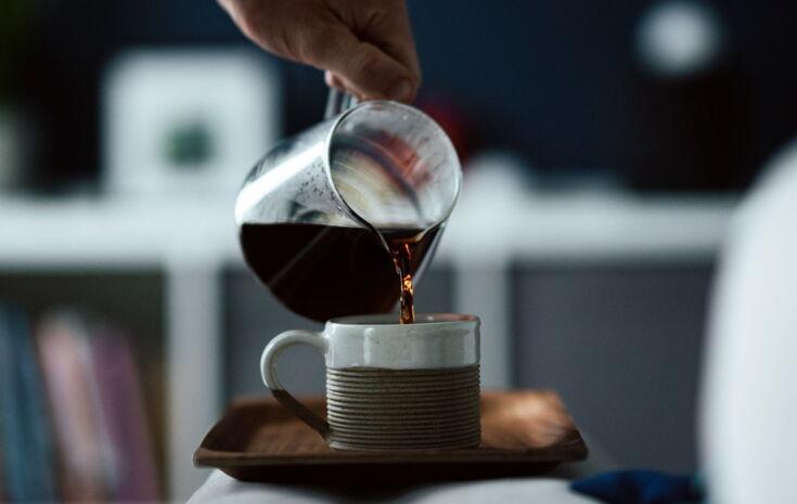 Person gießt schwarzen Kaffee aus Glaskanne in braunen Keramikbecher