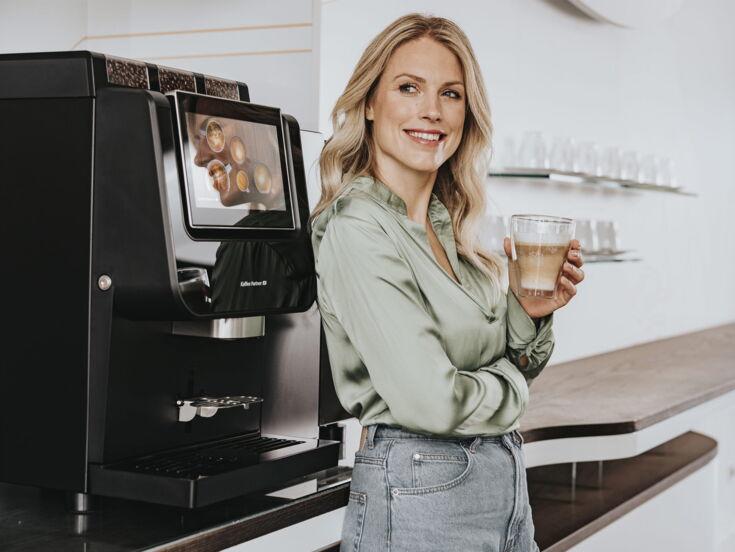 Eine lächelnde Frau lehnt an einem modernen Kaffeevollautomaten und hält ein Glas Latte Macchiato in der Hand in einem stilvollen Büro.