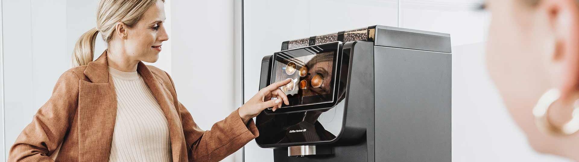 Eine Frau in eleganter Bürokleidung bedient einen Kaffeevollautomaten von Kaffee Partner im Büro. Eine weitere Frau ist im Vordergrund leicht unscharf zu sehen.