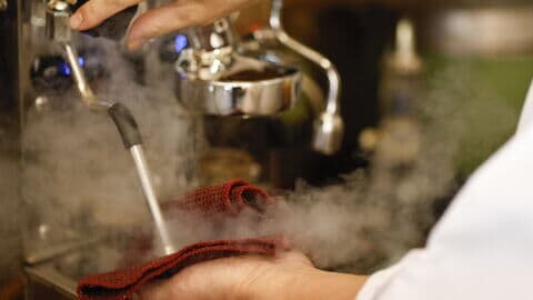 Eine Person reinigt die Milchaufschäumdüse eines Kaffeevollautomaten mit einem roten Tuch, während Dampf austritt.