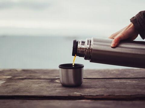 Eine Person gießt Kaffee aus einer silbernen Edelstahl-Isolierkanne in einen Becher auf einem alten Holztisch. Im Hintergrund ist eine ruhige, verschwommene Landschaft zu sehen.