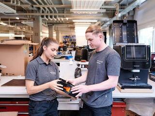 Zwei Servicetechniker, eine Frau und ein Mann, prüfen gemeinsam ein austauschbares Bauteil eines Kaffeevollautomaten in einer gut ausgestatteten Werkstatt.