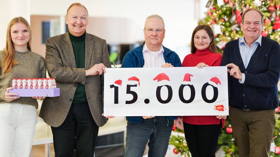 Fünf Personen stehen vor einem weihnachtlich geschmückten Hintergrund mit einem Weihnachtsbaum. Sie halten ein Schild mit der Aufschrift „15.000“, auf dem kleine Weihnachtsmützen dekorativ angebracht sind.