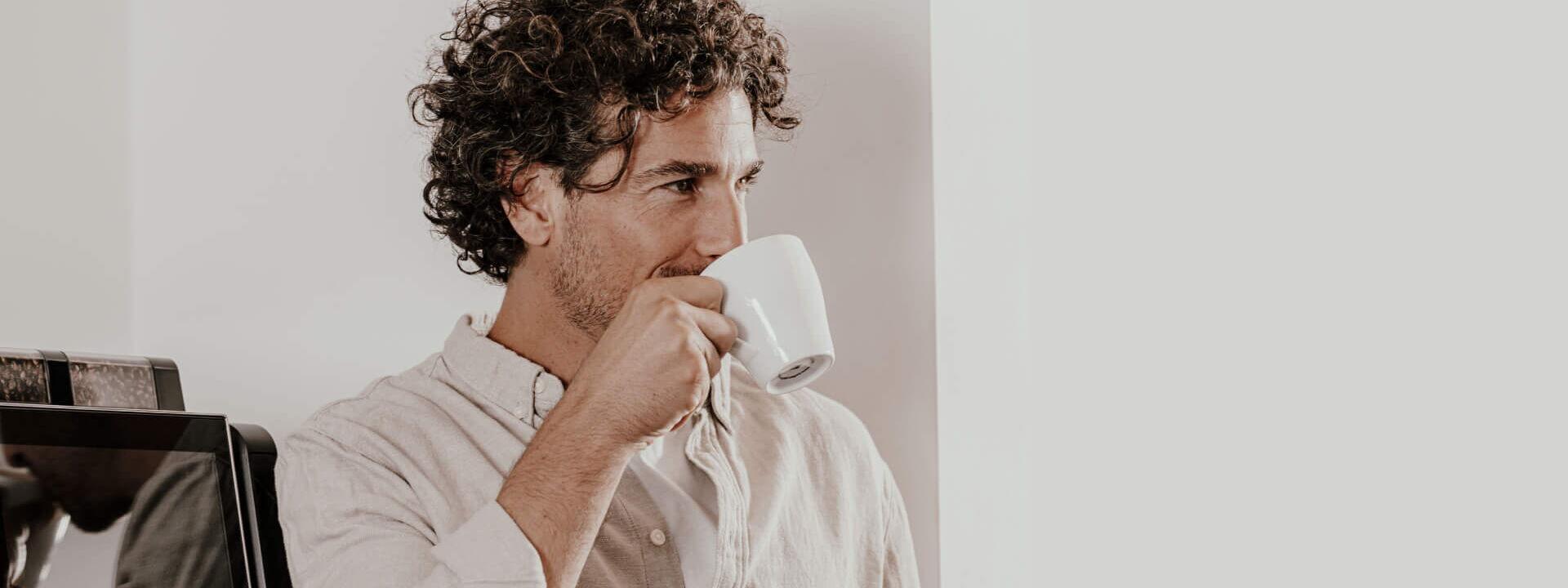 Ein Mann mit lockigem Haar genießt eine Tasse Kaffee, während er neben einem Kaffeevollautomaten von Kaffee Partner steht.
