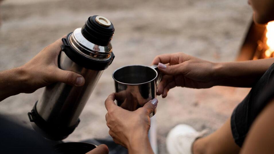 Person gießt Kaffee aus einer Thermoskanne in einen Becher bei einer Outdoor-Pause.