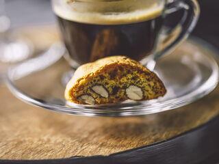 Ein halber Cantuccini Keks liegt als Beilage auf einer Untertasse, neben einer Tasse Kaffee