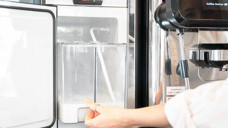 Nahaufnahme eines Kaffeevollautomaten von Kaffee Partner mit angeschlossenem Milchschlauch und Milchkühler, Hand zeigt auf den Milchkühler
