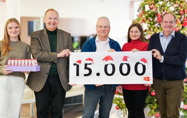 Fünf Personen stehen vor einem weihnachtlich geschmückten Hintergrund mit einem Weihnachtsbaum. Sie halten ein Schild mit der Aufschrift „15.000“, auf dem kleine Weihnachtsmützen dekorativ angebracht sind.