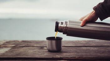 Eine Person gießt Kaffee aus einer silbernen Edelstahl-Isolierkanne in einen Becher auf einem alten Holztisch. Im Hintergrund ist eine ruhige, verschwommene Landschaft zu sehen.